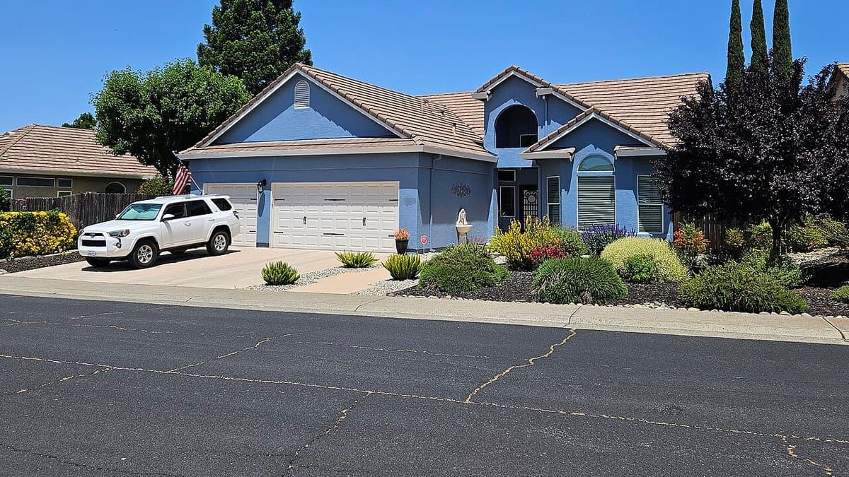A single-story blue house in Roseville, California with a front yard of succulents, gravel, and a small fountain in place of grass, and an SUV in the driveway
