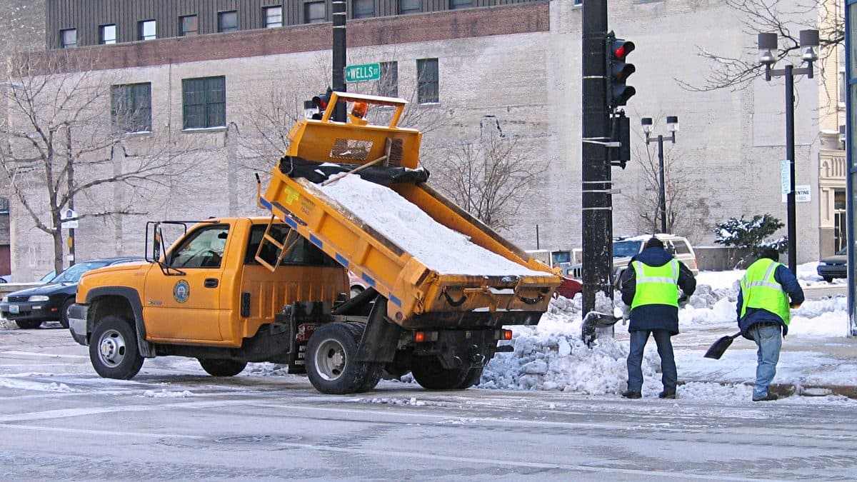 An orange city salt truck tipping a load of rock salt at a snowy street corner while two workers in safety vests shovel snow beside it