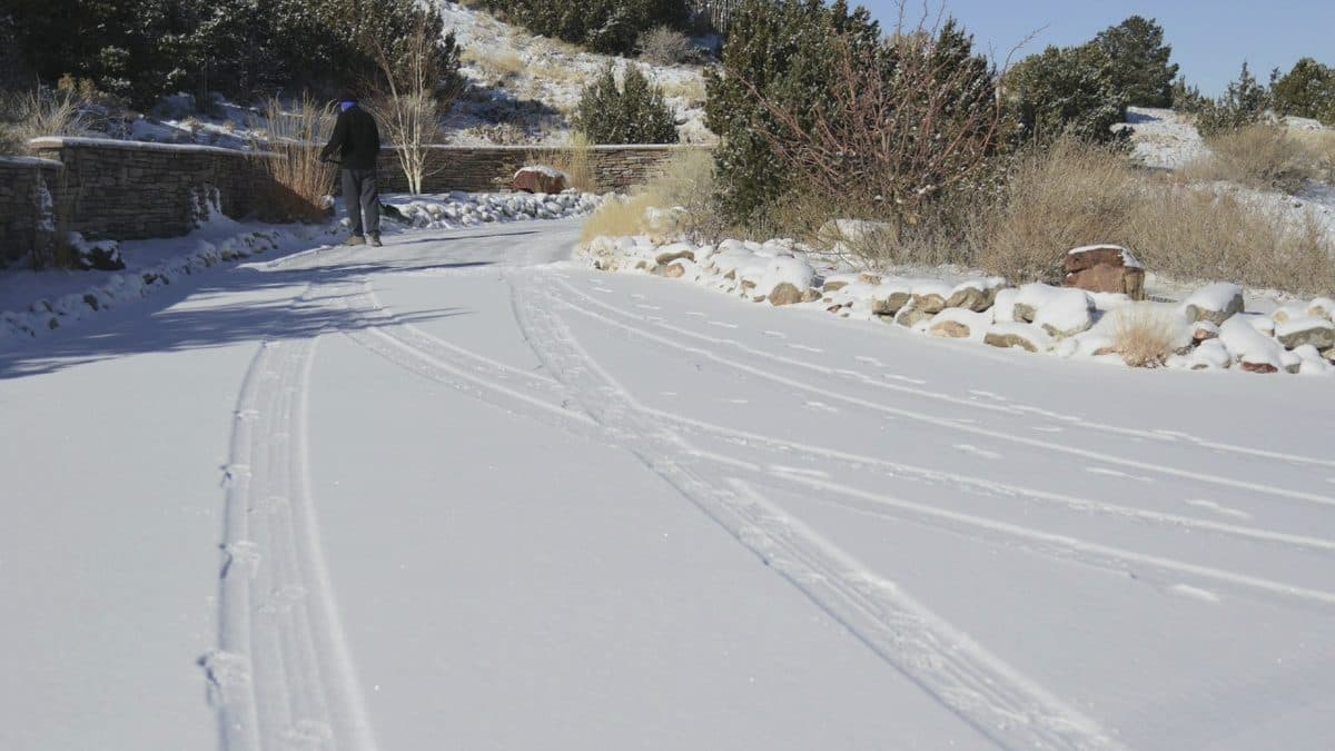 A person shoveling a long snow-covered driveway with tire tracks, edged by snow-capped boulders, shrubs and dry grasses under a blue sky