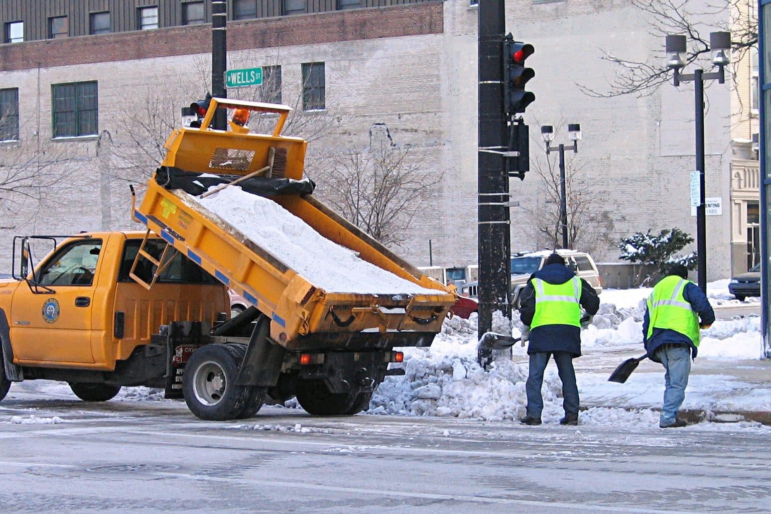 An orange city salt truck tipping a load of rock salt at a snowy street corner while two workers in safety vests shovel snow beside it