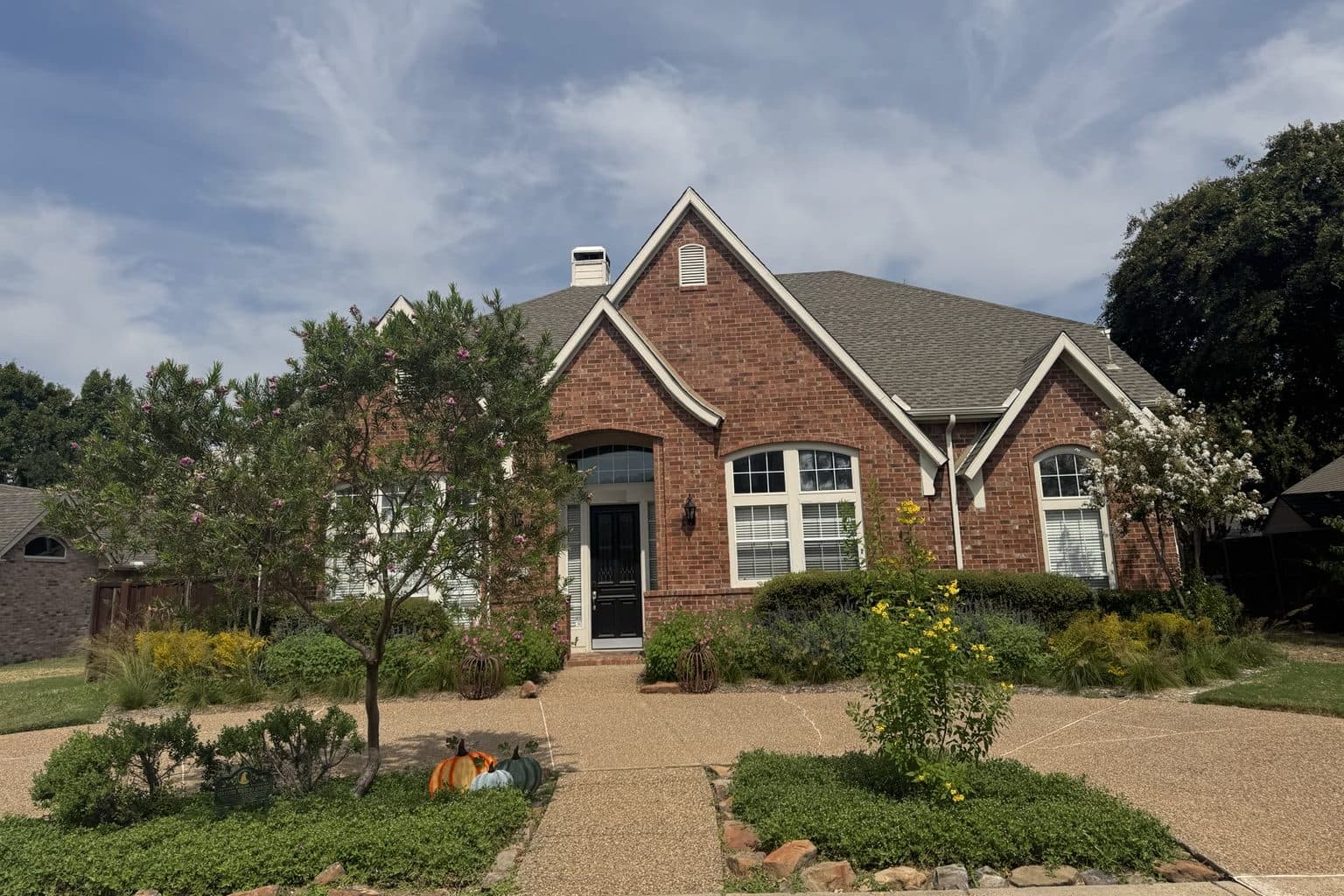 A brick house whose front lawn strip has been replaced with planting beds, with a desert willow and an esperanza in frogfruit-covered island beds