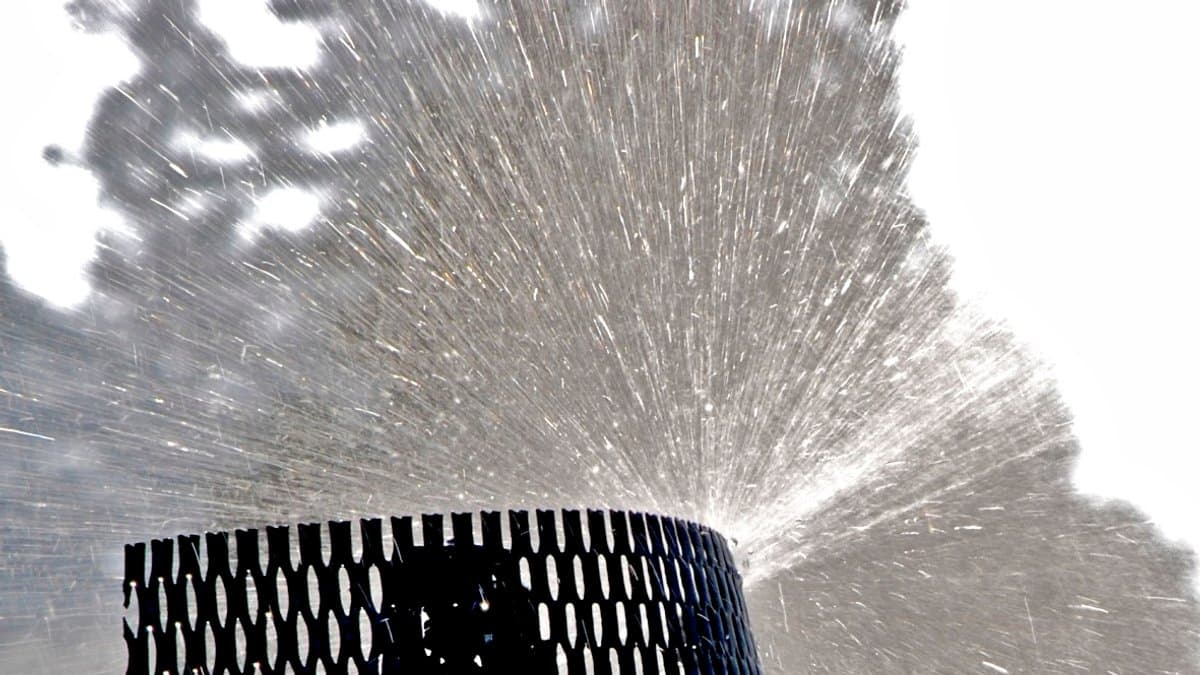 Close-up of a lawn sprinkler head spraying a wide fan of water droplets against a bright sky